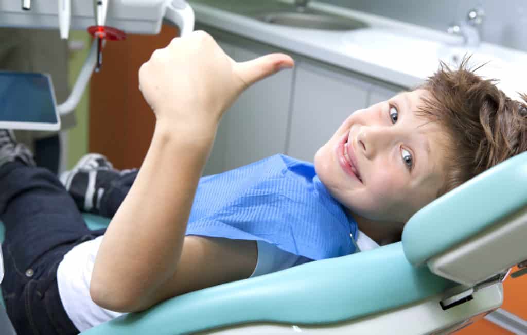 a young boy laying in a dentists chair smiling and giving a thumbs-up.