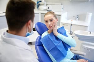 A blonde woman in a dentists chair conversing with her dentist while holding the side of her face in discomfort.